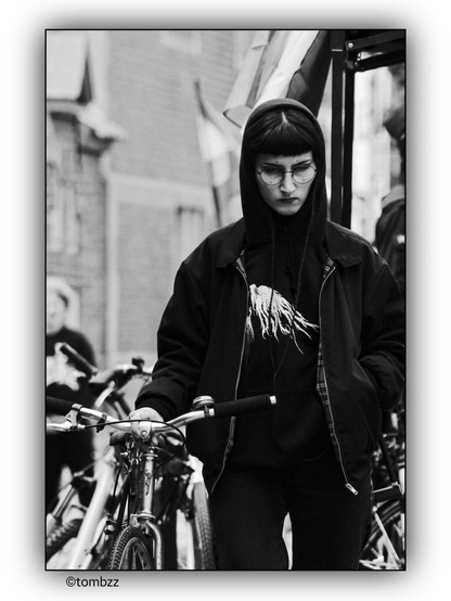 The black-and-white photograph captures a young woman walking her bicycle down the street. Her outfit draws attention—she wears a dark jacket and a hoodie with the hood up, covering her head. A graphic on her clothing, resembling tree roots, adds a unique touch to her overall appearance. She has a nose piercing, and her face is partially concealed by glasses, giving her a distinctive and intriguing look.

In the background, brick buildings emphasize the urban setting. The scene is dynamic—another person in motion appears in the frame, adding a sense of natural movement and liveliness. Several bicycles are visible on the street, suggesting that this area is frequently used by cyclists. The striking composition and details of the woman’s silhouette make her presence stand out within this environment.