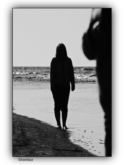 A black-and-white photograph shows a woman standing at the edge of the sea, her bare feet submerged in the water. She gazes toward the partial silhouette of a figure on the right side of the frame, which could be interpreted as a man. The stark contrast of light and shadow makes the image ambiguous, leaving ample room for interpretation and imagination.