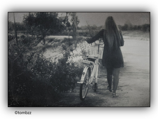 A woman with long blonde hair, wearing a dark dress, is walking with a white bicycle that has a basket attached to the handlebars. She is on a dirt road that curves to the left, with dense bushes visible on the left side. The analog photograph has noticeable grain, giving it a vintage character.