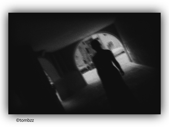 The photograph depicts the silhouette of a woman in a light dress, captured from behind, standing in a passage under an arch-like structure, resembling a gate or arcade. The composition is dynamic—the frame is tilted, adding a sense of unease and mystery. The black-and-white color scheme enhances contrasts and gives the photo a nostalgic, almost cinematic feel. In the background, slightly blurred buildings add depth and an urban atmosphere. The entire image has a subtly dramatic, almost poetic mood.