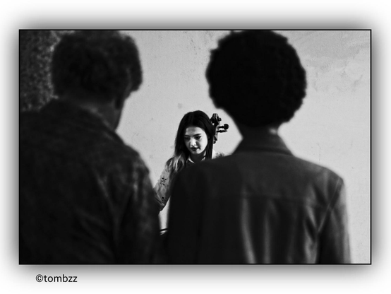 The girl at the center of the frame is playing the cello, leaning slightly over the instrument. Her long, dark hair cascades over her shoulders, and her focused expression highlights her complete immersion in the music. She holds the bow in her hand, gliding it harmoniously across the strings.
In the foreground, a couple—a woman and a man—both have distinctive afro hairstyles. Their silhouettes are blurred, making the girl the primary focal point of the image. The contrast between the sharpness of the central figure and the soft, indistinct outlines of the couple adds depth to the photograph. The black-and-white color scheme enhances the atmosphere of concentration, giving the image an elegant, artistic character.