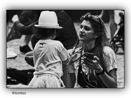 The girl stands facing her mother, dressed in a Puma-logo shirt and a light skirt. She wears a bright hat that contrasts with the darker background. The mother kneels before her daughter, holding a phone in one hand while gently smoothing a strand of her own hair with the other. Her lips are slightly parted, suggesting that she is speaking to the girl. In the background, pedestrians and a baby stroller add to the everyday urban atmosphere. The black-and-white color scheme enhances the emotional depth of the photograph, highlighting subtle details of their relationship and surroundings.