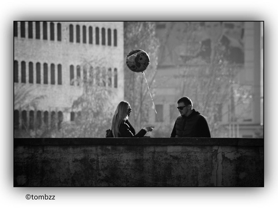 A black-and-white photograph captures a woman and a man standing on a bridge. Their conversation appears tense—the man has a serious expression, and the woman is pointing her finger at him, possibly emphasizing her words. Above them floats a helium-filled balloon, held by a string. The string is grasped by a young girl, their daughter, who remains out of sight behind the bridge's wall. The background is rich in detail and contrasts with the foreground, adding depth to the scene.