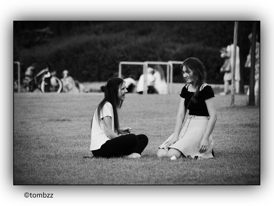 In the black-and-white photograph, two girls are sitting on the grass in the foreground. One has long, loose hair and is wearing a light-colored shirt with dark pants. The other has her hair tied in a braid and is dressed in a dark blouse and a light skirt. Both seem to be engaged in conversation or observing their surroundings. In the background, a group of people is visible—some sitting on benches, others standing or chatting. Several bicycles are leaned against park benches, suggesting that the place is popular among cyclists. The trees and greenery in the distance create a peaceful atmosphere of an urban park or recreational area.