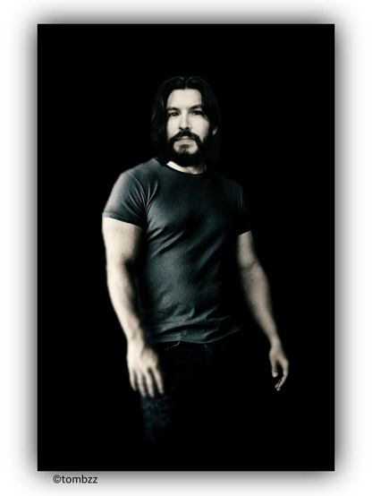 Portrait of a man against a black background. He is wearing a dark gray short-sleeved t-shirt that fits snugly, highlighting the muscular build of his arms and chest. His lower body is dressed in dark trousers. He stands upright with his arms relaxed at his sides.  

His face is sharp and expressive, with a well-defined jawline and high cheekbones. He has dark, thick eyebrows and deep-set eyes that appear focused. His nose is straight, and his lips are slightly parted. He sports a short, well-groomed beard along with a mustache, giving him a rugged yet elegant appearance.  

The overall portrait creates a strong, distinct image, accentuated by the contrast with the black background.