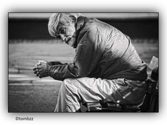 An elderly man sits on a wooden bench, leaning forward with his elbows resting on his knees and his hands clasped together. His gaze appears distant, as if lost in deep thought. He is dressed in a dark, slightly worn jacket and neutral-colored pants, suggesting practical attire suited to the weather conditions. On his feet, he wears shoes that seem somewhat worn. The background of the image is blurred, but it is clear that he is in a public space—possibly a park or a city square. The atmosphere encourages reflection on the history and situation of the man in the picture.