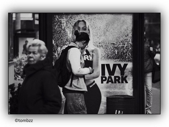 The photo captures a bus stop with an advertising billboard for the brand IVY PARK. In the foreground, there is a blurred figure of an elderly woman wearing a dark coat. In the background, to the left, a young girl with a backpack stands facing the billboard. The advertisement features a slender, young woman with a flawless physique, dressed in sportswear. The contrast between reality and the idealized image in the advertisement is striking—the girl appears to be confronted with the aspirational portrayal on the poster. On the right side of the image, there is likely a man also waiting for the bus. The photograph is in black and white, emphasizing its sharpness and the contrast between the elements in the scene.