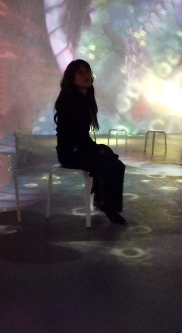 
Una mujer con cabello castaño oscuro y largo, sentada de perfil en una silla blanca sencilla en una habitación con paredes iluminadas con proyecciones de colores pastel.  El fondo es una mezcla de formas y colores difusos, que parecen proyecciones abstractas. La mujer viste de negro. Hay otras sillas vacías de metal en el fondo.  La escena tiene un ambiente misterioso y algo surrealista.