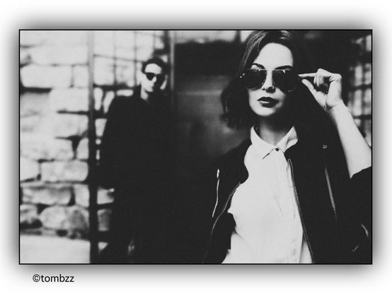 A black-and-white photograph features a woman in the foreground and a man in the background. The woman is wearing a light-colored shirt and a dark zip-up jacket. She is making a gesture with her hand as if adjusting her sunglasses. The man in the background is also wearing sunglasses and is dressed in dark clothing. Behind them, there is a stone wall and a metal grate, adding to the mysterious and enigmatic atmosphere of the image. The composition is thoughtful, suggesting a story or a relationship between the two figures.
