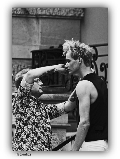The photo shows two young men. One of them is wearing a floral-patterned shirt, and both of his forearms are adorned with tattoos. He is clearly shorter than the other man, who is dressed in a black vest and white trousers. He is cosmetically adjusting his face before a photo session. The background features a section of a building with stone elements and a metal railing, giving the scene a raw, industrial character. The photo is in black and white, which accentuates the contrasts and details of the composition.