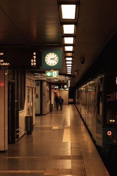 A modern train station bathed in warm, golden light. A silver train with red accents is parked at the platform. A few travelers walk by, some with luggage. The platform is clean and well-lit, with clear signage.