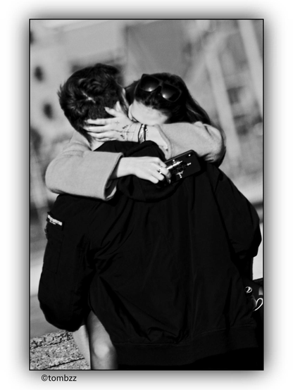 A black-and-white photo captures a loving couple—a young man and woman—closely embracing each other in a street setting. The woman has sunglasses pushed up onto her hair. Her hand rests on his neck, while in her other hand, which bears a visible tattoo, she holds a smartphone. The warmth and affection between them are evident in every gesture.