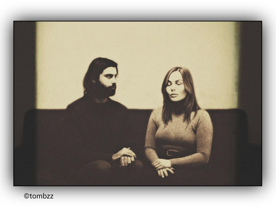 A monochrome analog photograph depicts a couple sitting on a classic sofa, reminiscent of traditional marriage portraits. The man, dressed in a dark sweater and pants, sits slightly turned toward the woman. He looks at her as if trying to grasp her presence, while she, dressed in a light sweater cinched with a belt, has her eyes closed. She is there, yet not quite.
The positioning of their hands emphasizes the tension in the scene—his fingers intertwined, seemingly holding back emotion, while her hands rest on her lap, distant and enclosed in her own world. The conventional portrait style is deliberately broken—rather than harmony, there is a subtle fracture in the relationship, an underlying narrative of presence and absence.
The photograph has a deep tonal structure, where light and shadow create an added emotional dimension. In the background, darker, scattered stripes appear, adding a nostalgic character—as if the image not only captures a moment but also preserves the fleeting nature of memories. It references the style of vintage photographs but gives them a new meaning, transcending the boundaries of traditional representation.