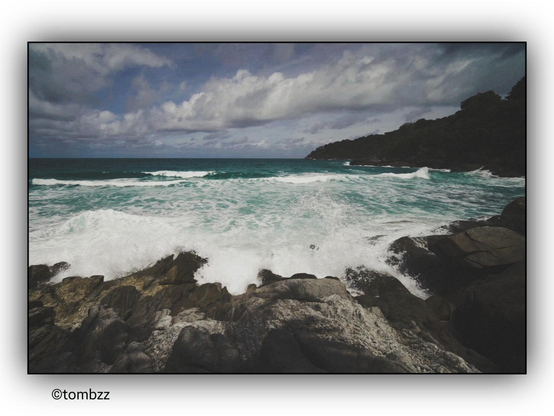 The image depicts a picturesque seascape. In the foreground, there is a rocky coastline where waves crash against the shore. The water is turquoise, with foamy white waves. In the background, the horizon stretches across a deep blue sea, with clouds floating above. On the right side of the image, forested hills descend toward the water. Altogether, the scene presents a tranquil, natural landscape.