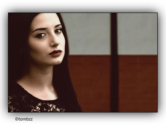 Portrait of a young woman with dark hair and striking lips. Her gaze is intense, full of depth, as if she holds a secret. She wears a black lace blouse, adding a touch of subtle elegance to her appearance. The background is divided into two sections – the upper part is light, while the lower part is a deep, intense red, creating contrast and emphasizing the air of mystery. A vertical dark stripe runs through the background, adding dynamism to the composition. The overall effect is captivating, with a dramatic atmosphere and a carefully balanced interplay of light.