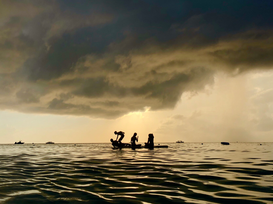 Photo of a beach raft in the middle distance, with four teenagers silhouetted against the golden glow of the coming sunset, heavy clouds casting a shadow on the scene  ￼