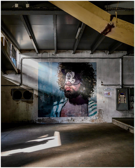 A large mural of a man with curly hair and facial paint, resembling a clown, is featured on a grungy wall in an industrial space. Sunlight streams in, casting shadows on the concrete floor. The surroundings include hanging pipes and air conditioning.
Photo taken during the Ibug exhibition in August 2024