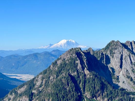 Mount Rainier from the Alpine Lakes section of the PCT. Snow topped mountain with rocky mountains in the foreground. Light blue clear sky.