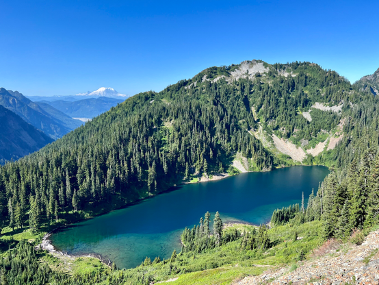 Joe Lake along the Alpine Lakes section of the PCT. Greenish alpine lake at around 5,500 feet. Trees surround part of the lake. Mount Rainier in the distance. Pretty blue sky.