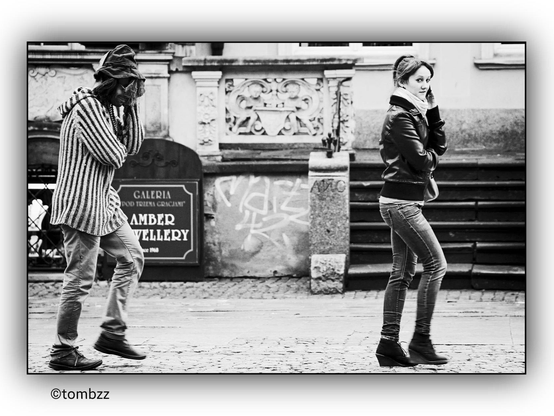 On the street, a young woman is talking on the phone, holding her smartphone to her ear. Behind her, a man follows her steps, mimicking her movements, especially her gestures and the way she holds her phone. The woman is wearing a leather jacket, jeans, and boots, while the man is dressed in a striped sweater, light-colored pants, and a hat. The background features an urban setting with cobblestone pavement and a graffiti-covered wall, enhancing the street photography character. The photographer captured the moment when the woman realized she was being photographed, but she was unaware of the person behind her.
