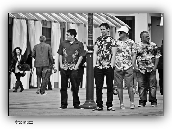 Four guys standing in the street, rocking tropical shirts covered in palm trees, looking like they just stumbled out of a legendary all-night party. Hands shoved deep in their pockets, as if holding onto the last bits of dignity. Their expressions? A mix of exhaustion, disbelief, and maybe even silent protest against the morning sun. They totally give off "The Hangover" vibes—like a crew desperately trying to piece together the events of last night. In the background, people walk by, oblivious to the drama, while white pavilion curtains and city buildings frame this unforgettable scene.