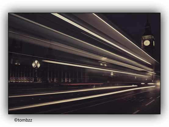 The photo captures a nighttime scene featuring Big Ben in London, taken using a long exposure technique.** This allows passing vehicles to leave dynamic light trails stretching across the width of the frame. The red and white lights create a sense of movement, contrasting with the still architecture of the city. In the background, the well-lit tower of Big Ben stands majestically, with its clock clearly visible. In the foreground, streetlights and sections of buildings add depth to the composition. The road displays the words "BUS LANE," slightly blurred due to the long exposure time.
Thanks to this technique, the photograph gains a dynamic quality, showcasing street traffic as striking streaks of light while the historic building remains a static focal point in the nighttime London scenery