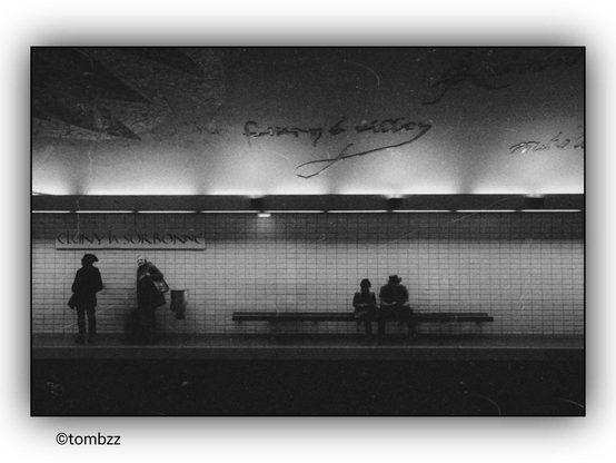 The image shows the Cluny La Sorbonne metro station. In the foreground, four people are visible: two standing on the left side and two sitting on a bench to the right. The photograph is in black and white, giving it a nostalgic and artistic feel. In the background, the wall features the inscription "CLUNY LA SORBONNE".
