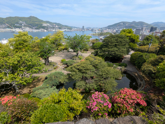 Büsche und Bäume in einem Park in der Vogelperspektive.
Im Hintergrund der Hafenbereich und die umliegenden Berge von Nagasaki. 
Vorne rechts zwei Büsche mit pinken Blüten. 