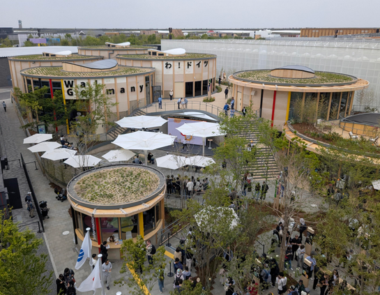 Blick auf die begrünten und aus hellem Holz bestehenden, kreisrunden Deutschland Pavillon.
6 Gebäude. Dazwischen sind weisse Schirme aufgespannt. 