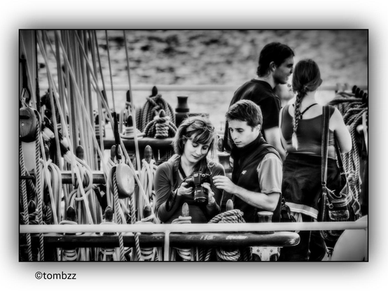 A black-and-white photograph captures four people on the deck of a sailing ship. The main subjects are a young woman and a man leaning over a camera. The woman holds the camera in her hands, browsing through the photos with a focused expression. The man, standing close to her, looks at the pictures with interest and extends his hand as if pointing out something important. His face conveys curiosity and concentration, while the woman has a slightly smiling, satisfied expression.  
The young woman is wearing a light summer dress, perfect for a day at sea, and her hair is loosely flowing. The man next to her is dressed in a short-sleeved T-shirt and a sporty vest, emphasizing his functional and casual style suited for conditions on board. In the background, another couple is engaged in conversation. The woman wears a sleeveless top and a skirt, while the man wears a short-sleeved T-shirt.  
The setting clearly indicates that they are on a sailing vessel—ropes, masts, winches, and rigging elements are visible. In the background, the sea stretches beyond the ship’s railing.