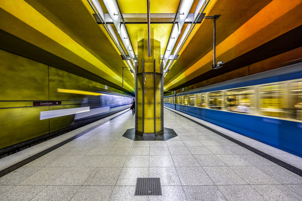 Symmetrical shot of the platform at Candidatz metro station in Munich, Germany with a dominance of greenish yellow colors on the wall and a bit of orange. Two blue metro train are visible on each side of the platform. One is a modern type one and is speeding up, fading away in the distance, revealing the wall and nameplate of the station. Three splashes of light are also pointing on the direction of the silhouette of a passenger walking on the platform. The other train is from an older type and arriving slowly, breaking down on the platform and revealing passenger silhouettes.