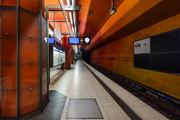 Side view of the plaftorm at Candidatz metro station in Munich, Germany, with a dominance of dark oranges tones. A big orange pillar is visible on the left side in the foreground.