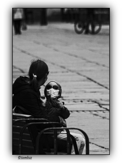 A black-and-white photo shows a mother sitting on a bench with her child on her lap during the COVID-19 pandemic. The mother is wearing a dark jacket, sunglasses, and a protective face mask. Her hair is tied in a ponytail. The child, also wearing a mask, is dressed in a dark hooded jacket, has a cap on their head, and is wearing sunglasses. The background features a section of the sidewalk with visible lines and cracks, and in the distance, a blurred silhouette of a person and a bicycle can be seen.