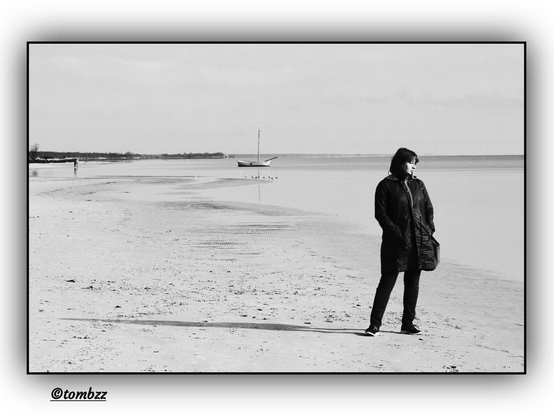 Seaside beach. In the foreground, a woman dressed in dark pants and a jacket, with her eyes closed, possibly lost in thought. Her silhouette contrasts distinctly with the surroundings. In the background, a sailboat and, to the left, two people. A black-and-white photograph capturing the tranquility of the scene, with the smooth surface of the water in the bay.