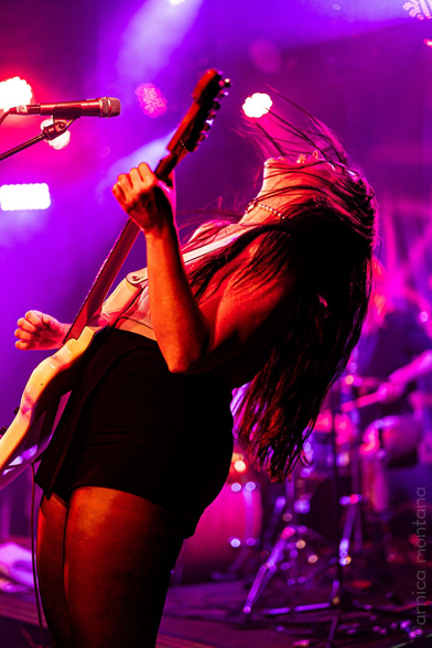 A musician passionately plays an electric guitar on stage, head tilted back, with long hair flowing. The scene is illuminated by vibrant purple and red lights, creating an energetic atmosphere. In the background, blurred instruments are visible, indicating a live performance setting.