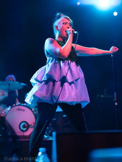 A singer performs on stage, wearing a sleeveless, colorful dress and holding a microphone. The scene is illuminated by blue stage lights, and a drummer is visible in the background.
