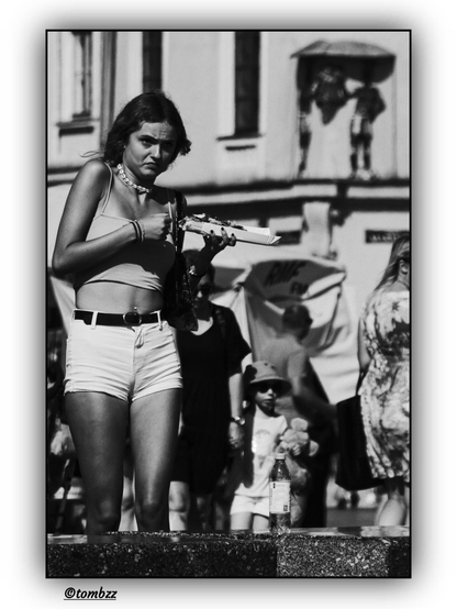 A black-and-white street photograph captures a young girl holding Polish street food. She is wearing a sleeveless crop top and high-waisted shorts, tied with a black belt. A delicate necklace hangs around her neck, and she has a bracelet on her wrist. Her facial expression reveals nervousness—a small wasp is circling around her, affecting her posture and demeanor. In the background, pedestrians can be seen, including a child holding a plush toy. A bottle of water rests on the ledge nearby. Further back, there is a building with windows and an umbrella under which two people are standing. The whole scene is filled with the energy of street life, while the black-and-white aesthetic adds a nostalgic and artistic feel.