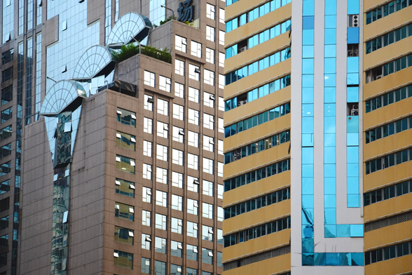 A composition of several skyscrapers next to each other with a variety of wallfaces in different directions and colours. To the right a yellow building looks to the left, it has horizontal yellow levels with dark cyan windows. Horizontally a white blue striped outset view to the right, to the camera. To the left of the building a different one outstretches far to the images side. It is staggered with half round glass arches ontop. The right facing side has a grid of white windows and dark-sand coloured stones. It’s left showing face has a large wall of glass, framed by vertical window stripes 