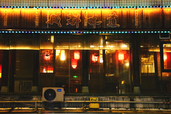 A photo of a window front with a worn out look. Inside of the dirty windows are red lanterns, they glow dimly. Ontop yellow LEDs illuminate a worn out sign with a few Chinese characters on it. One AC unit decorates the outside. It’s a moody dark, yet colourful image 