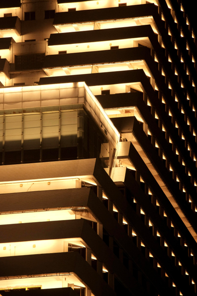 A photo of a tiled, ledge structure. It seems to be a building with many floors and a staggered front. It seems to have balconies all around with yellow lights in them. The outside is rather dark, it seems to be nighttime. The balconies and a blocky outset in the front give off a strong glow 