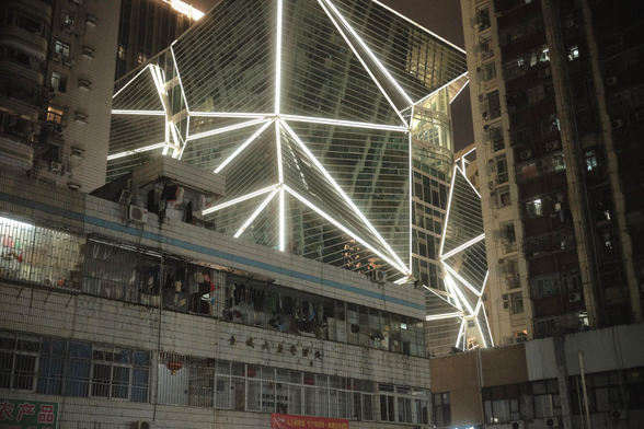 A photo of three living complexes surrounding three sides of the image. In the middle, behind the living quarters rests a rather modern building with walls in arbitrary angles, cut into triangles. lined with whitw lights. Its a stark contrast  