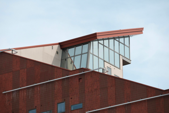 A photo up a red brick building with leading lines in non typical angles. One roof-line rises from the right to the left, leaving the image, but coming back and rising higher back to the right, ending in a sharp angled roof with a large wall of windows. It’s an intriguing scene. The bottom half of the image is a red brick wall with a few windows, the top half is grey-blue sky 