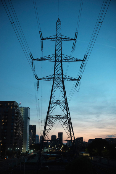 A tall power line under a teal evening sky. Some buildings are visible to the left and far ground. The powerlines leave the image to the top, the sky is lightly textured 