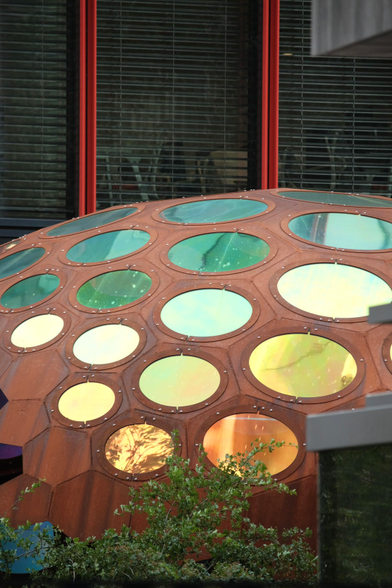 A photo of a half sphere with hexagonal faces. Each face, all of different sizes, features a round colored window. The colours seem to be metallic, having a weirdly narrow spectral glow to them. Behind the sphere a building with large windows, closed metallic blinds and red metallic bars grows 