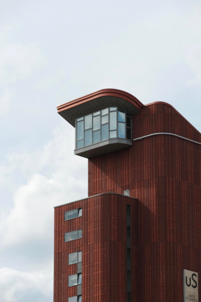 A photo up a brown-red building. It has a weird shape for a building, chamfered corners and a lightly sloped roof to the right. To the left of the building there is no sloped roof but an outset rounded room with a staggered array of windows. It’s a strange scene 