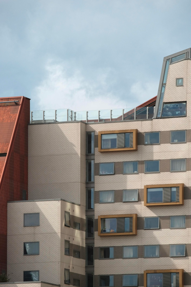 A photo of a building structure that is very non typical. To the left a brown sharp angled roof rises up. To the right of it a beige brick building emerges, it has multiple faces and a roof with various angles. Some yellow outset windows attract the viewers attention 
