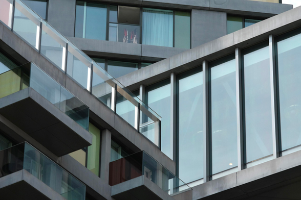 A photo of several living quarters in a composed fashion. To the top a high rise is almost parallel to the camera. It is grey, down to the right a walkway leaves the image at an angle, with vertical large windows. To the left below that another high rise leaves the image in the opposite direction. It has colo ur ed faces and glass balconies, but is still grey 