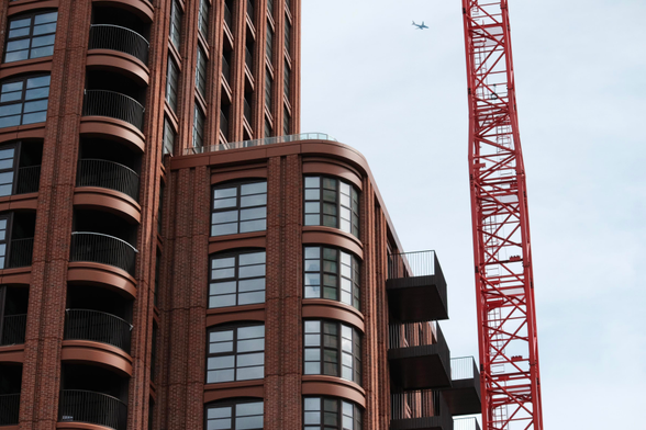 Photo of a brown-red brick house with rounded edges and steel-silver windows. A crane enters and leaves the image to the right, a sole plane is visible in the sky 