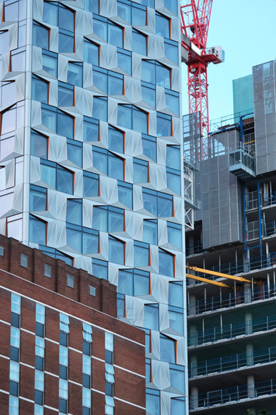 A collection of several high rises next to each other, most prominent is the center building, a white blue one with an elaborate window pattern and red accents. To its left and much less tall a red brick building rises. To its left a high rise under construction is visible. It is cluttered in building materials. Yellow fixtures fix it to seemingly the white building. A red crane grows into the sky 