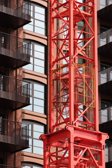 A photo up a red brick building with apartments stacked onto each other. Center on the wall are two columns of windows. To their sides balconies with brown fencing. Center blocking the windows is a red painted crane with some yellow caging 